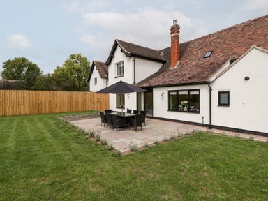 A backyard with a patio table and chairs under an umbrella next to a white house with a tiled roof and wooden fence at Byways in Madresfield near Malvern