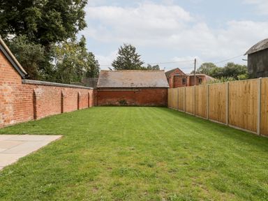 A grassy backyard enclosed by a wooden fence and a brick wall with a small brick building at the back at Byways Madresfield near Malvern