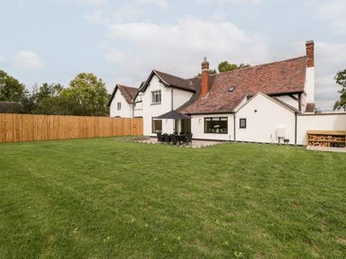 A large grassy backyard with a patio set and umbrella next to a white house with a tiled roof and wooden fence at Byways in Madresfield near Malvern