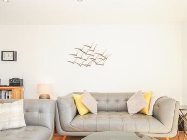 A living room with gray sofas with cushions a wall clock and a wooden cabinet with a radio at Pebbles in Downderry