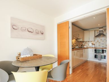 A dining area with a round table and four chairs next to an open doorway leading to a kitchen at Pebbles in Downderry
