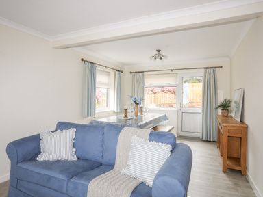 A living room with a blue sofa pillows and a blanket a dining table with chairs flowers and candles and a wooden cabinet with plants and a picture at 7 Maes Awel in Abersoch