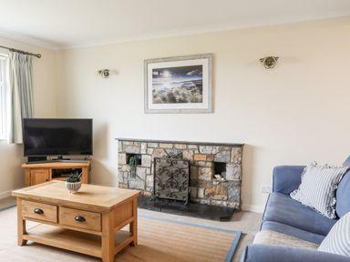 A living room with a wooden coffee table a television on a wooden stand a stone fireplace and a blue sofa at 7 Maes Awel in Abersoch