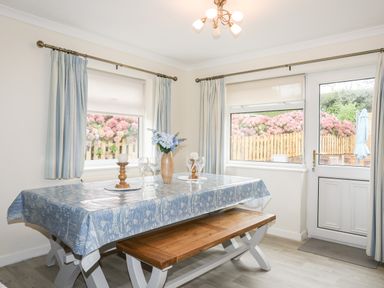 A dining area with a wooden bench table covered with a blue tablecloth and set with glasses and a vase of flowers at 7 Maes Awel in Abersoch