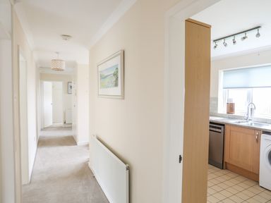 A hallway with beige walls and carpet leading to multiple doors and a kitchen with wooden cabinets and tiled floor at 7 Maes Awel in Abersoch
