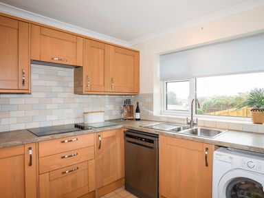 A kitchen with wooden cabinets a dishwasher a washing machine a double sink and a window at 7 Maes Awel in Abersoch