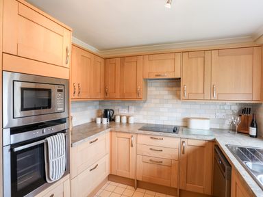 A kitchen with wooden cabinets a microwave oven countertop kettle mugs chopping board knife set wine bottle and sink at 7 Maes Awel in Abersoch