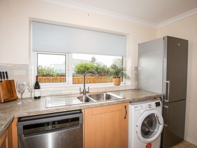 A kitchen counter with a double sink dishwasher washing machine refrigerator wine bottle wine glasses and a plant on the windowsill at 7 Maes Awel in Abersoch