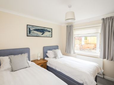 A bedroom with two single beds with striped pillows a wooden bedside table with a lamp striped curtains a window and a whale painting at 7 Maes Awel in Abersoch