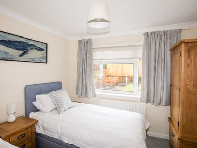 A bedroom with a single bed a wooden nightstand with lamp a wooden wardrobe and a window with striped curtains at 7 Maes Awel in Abersoch