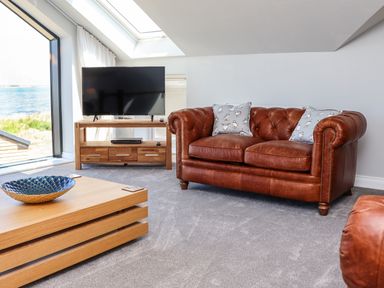 A living room with a brown leather sofa, a wooden coffee table, a television on a wooden stand, and a large window showing a sea view at The Waves in Amble