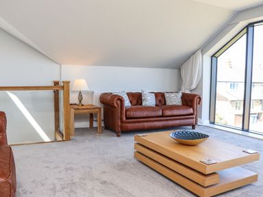 A sitting area with a brown leather sofa a wooden coffee table with a decorative bowl and a side table with a lamp by a large window at The Waves in Amble