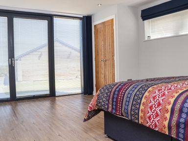 A bedroom with a patterned bedcover a wooden door and large glass doors with blinds at The Waves in Amble