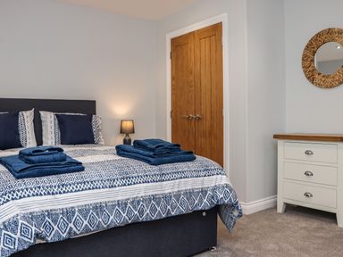 A bedroom with a bed with blue and white bedding and towels on it a lamp wooden double doors and a white chest of drawers with a round mirror at The Waves in Amble