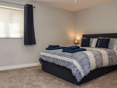 A bedroom with a bed with patterned bedding two pillows two sets of folded towels a window with blinds and dark curtains and a bedside lamp at The Waves in Amble