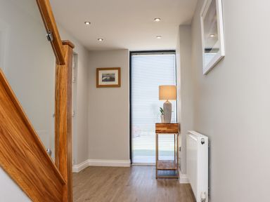 A hallway with wooden stairs a window with blinds a side table with a lamp and a plant a radiator and framed pictures on the walls at The Waves in Amble