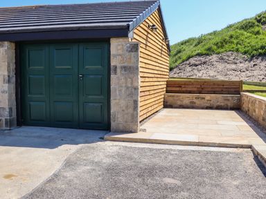 A garage with green doors next to a paved area and a grassy hill at The Waves in Amble