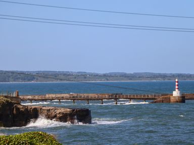 A pier with a red and white striped lighthouse extending into the sea with waves hitting rocks at The Waves in Amble