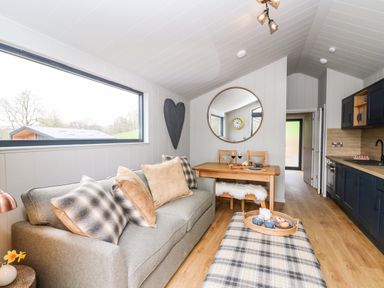 A living room with a sofa and cushions a wooden dining table with chairs a large round mirror and a kitchen area at Fawn's Rest in Nantmel near Rhayader