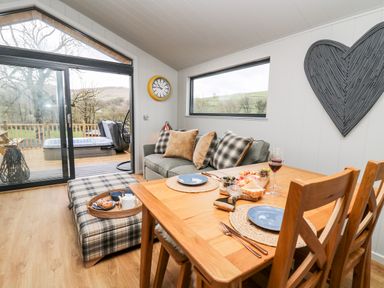 A dining table set for two with plates and wine next to a sofa with cushions and an ottoman in a room with large windows and a sliding glass door at Fawn's Rest in Nantmel near Rhayader