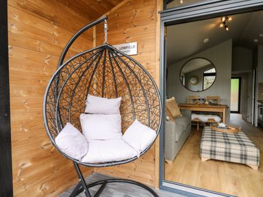 A hanging chair with cushions on a wooden porch beside a living room with a sofa and a plaid ottoman at Fawn's Rest in Nantmel near Rhayader