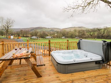 A wooden deck with a picnic table holding wine glasses and snacks next to a jacuzzi overlooking green fields and hills at Doe Lodge in Nantmel near Rhayader
