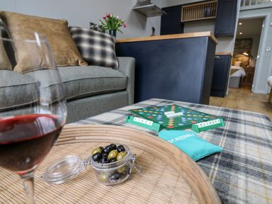 A living room with a sofa and pillows a tray with olives and a glass of red wine a Scrabble game on a plaid ottoman with a kitchen and bedroom visible at Doe Lodge in Nantmel near Rhayader