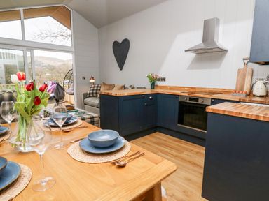 A kitchen with blue cabinets and wooden countertops next to a dining table set with plates and glasses and a vase of flowers at Doe Lodge in Nantmel near Rhayader