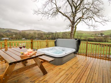 A wooden deck with a picnic table and a hot tub overlooking green fields and hills at Doe Lodge in Nantmel near Rhayader