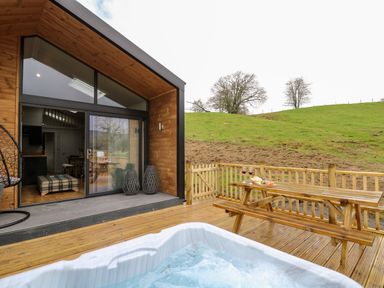 A wooden deck with a hot tub and picnic table next to a modern cabin and grassy hill at Doe Lodge in Nantmel near Rhayader
