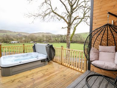 An outdoor wooden deck with a hot tub and a hanging chair with cushions at Doe Lodge in Nantmel near Rhayader