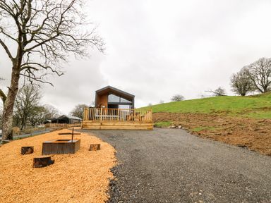 A small modern cabin with a wooden deck and fire pit area on gravel and wood chips next to grassy hillside at Doe Lodge in Nantmel near Rhayader
