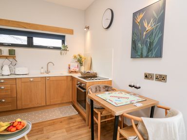 A kitchen with wooden cabinets a table with a Monopoly game two chairs a fruit bowl and a wall clock at Tin Shed Nantmel near Rhayader