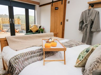 A bedroom with a bed holding a breakfast tray with orange juice and pancakes a bathrobe hanging on the wall and a chair by the window at Tin Shed in Nantmel near Rhayader