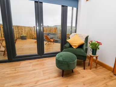 A green armchair with yellow pillow and ottoman next to a small wooden table with flowers near glass doors opening to a wooden patio at Tin Shed in Nantmel near Rhayader
