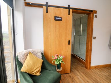 An armchair with a yellow pillow and a throw next to a sliding wooden door leading to a bathroom at Tin Shed Nantmel near Rhayader