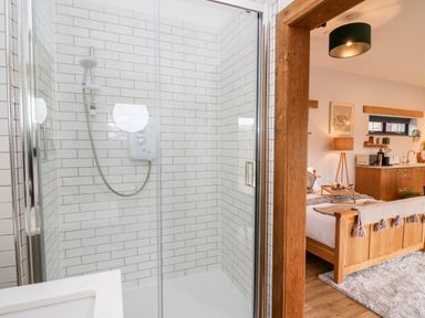 A shower with glass doors and white tiles next to a bedroom with a wooden bed and night lamp at Tin Shed in Nantmel near Rhayader