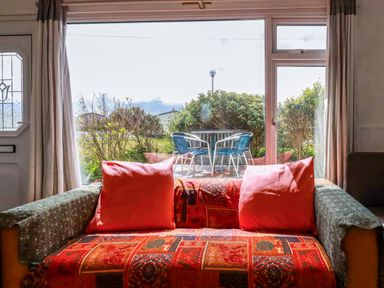 A sofa with cushions in front of a large window overlooking a patio with table and chairs at Sandpiper in Perranporth