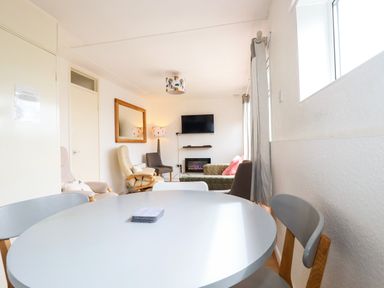 A dining area with chairs and round table leading to a living room with armchairs a sofa a wall mirror and a mounted tv at Sandpiper in Perranporth