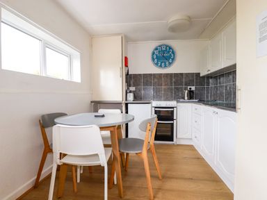 A kitchen with a round table surrounded by four chairs a stove a kettle and cabinets with a clock on the wall at Sandpiper in Perranporth