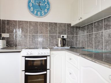 A kitchen corner with a white stove, kettle, toaster, and grey tiled walls at Sandpiper in Perranporth