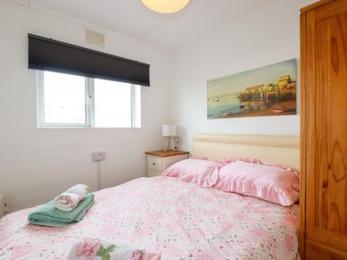 A bedroom with a bed covered in pink bedding a bedside table with a lamp wooden wardrobe and a window with a black blind at Sandpiper in Perranporth