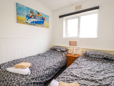 A bedroom with two single beds with patterned bedding a bedside table with a lamp and a window above the table at Sandpiper in Perranporth