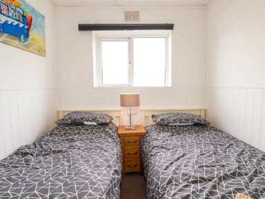 A bedroom with two single beds with geometric patterned bedding and a wooden nightstand with a lamp under a window at Sandpiper in Perranporth