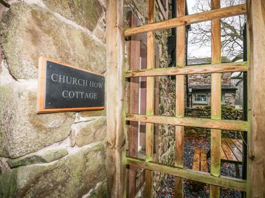 Stone wall with Church How Cottage sign wooden lattice gate outside a stone building with picnic table and leaf-covered ground at Church How Cottage in Nether Wasdale