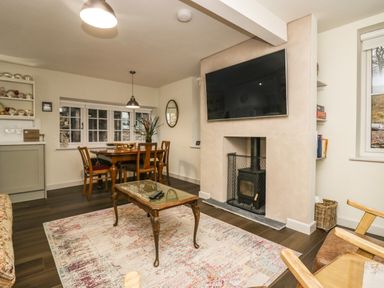 A living room with a dining table and chairs a coffee table a fireplace with a television mounted above and shelves with dishes at Church How Cottage in Nether Wasdale
