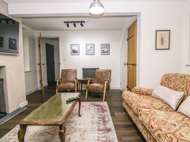 A living room with a patterned sofa two brown chairs a glass coffee table and framed pictures on the wall at Church How Cottage in Nether Wasdale