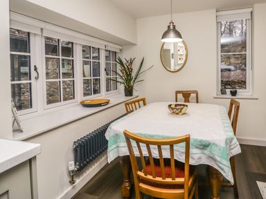 A dining area with a table covered by a tablecloth and wooden chairs near windows and a mirror at Church How Cottage in Nether Wasdale