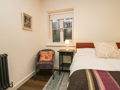 A bedroom with a bed with patterned pillows a chair with a cushion a window and a nightstand with a lamp at Church How Cottage in Nether Wasdale