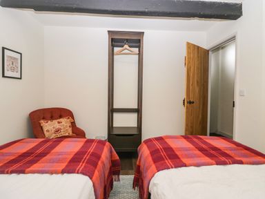 A bedroom with two single beds covered with red and orange plaid blankets a red chair with a pillow a wooden open wardrobe and a wooden door at Church How Cottage in Nether Wasdale